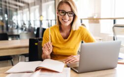 Photo of beautiful happy woman looking at camera while sitting Photo of beautiful happy woman looking at camera while working and sitting at table in open-plan office