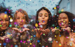 Young women blowing confetti from hands. Friends celebrating outdoors in evening at a terrace. Young women blowing confetti from hands. Friends celebrating outdoors in evening at a terrace.